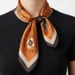 A female model wearing a black T-shirt and a square silk satin scarf in warm amber and brown tones. The scarf features an intricate compass-inspired  pattern with fine stripe accents along the edges, held stylishly in place around her neck with a dark brown leather scarf ring.
