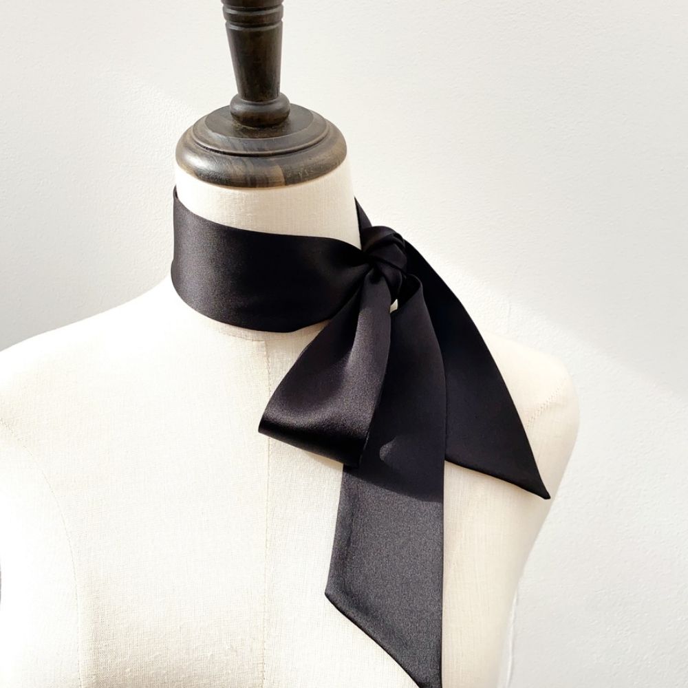 A long black mulberry silk satin skinny scarf tied in a large bow around the mannequin's neck
