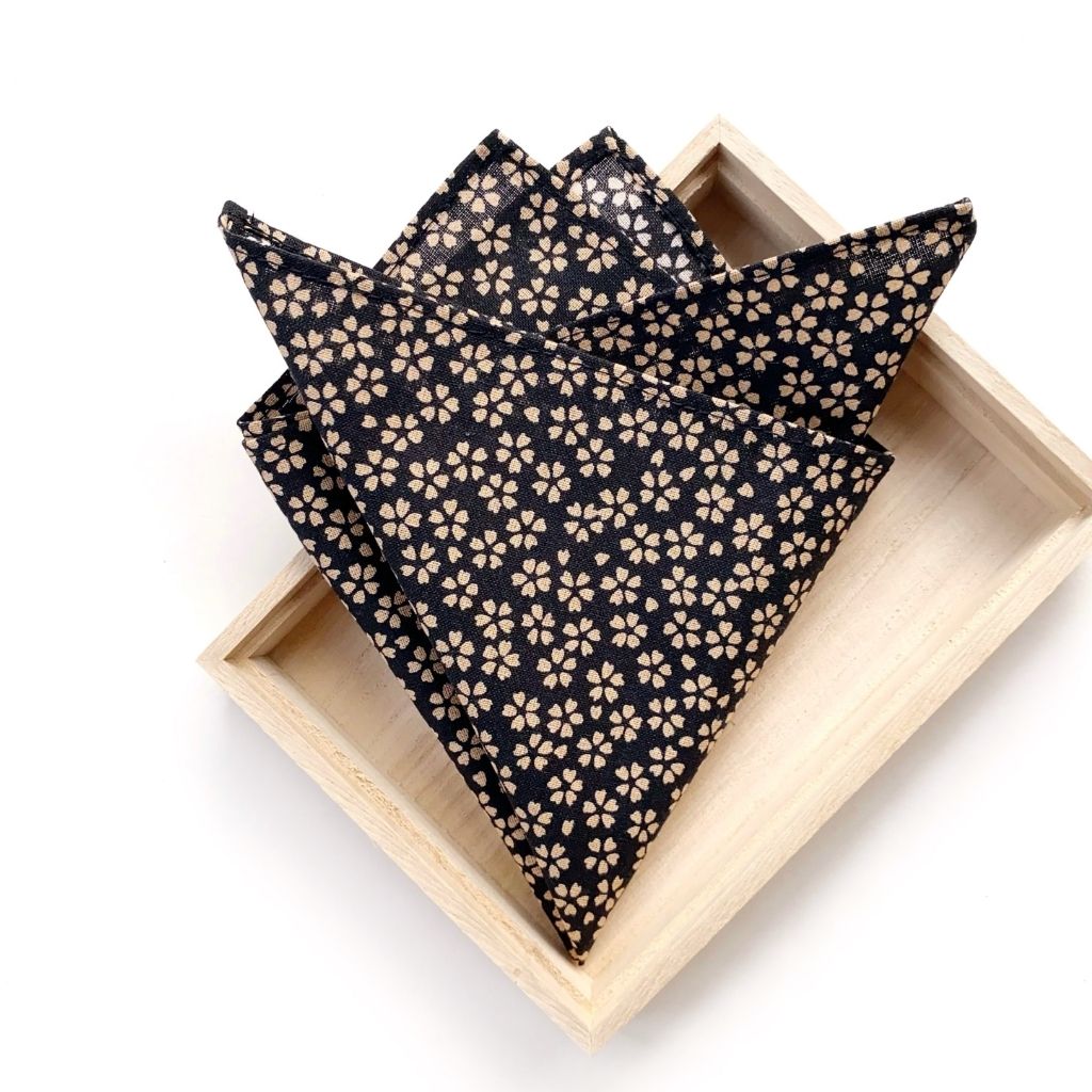 folded blak floral print japanese cotton pocket square hankerchief displayed on a light wood tray against a white background.