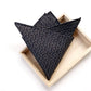 A folded black japanese cotton fabric pocket square with a repeating white wave and dot pattern, displayed on a light wood tray against a white background.