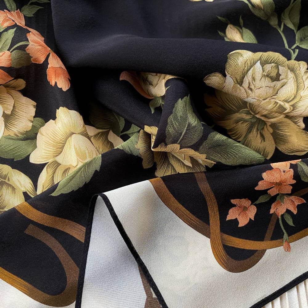 A close-up of a draped mulberry silk crepe de chine scarf featuring a dark navy background with large cream and beige flowers, green leaves, and peach-orange blossoms, plus metallic gold curved accents.