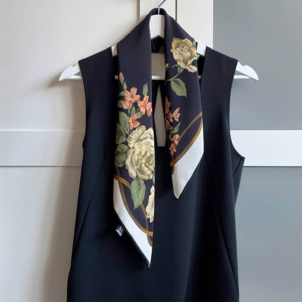 A black mulberry silk scarf with a floral print of cream roses, orange flowers, and green leaves, draped over a sleeveless black dress on a white hanger.