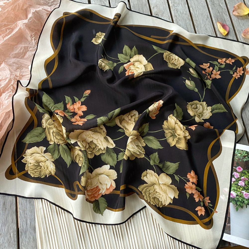 A square mulberry silk scarf with a black background, featuring cream and peach roses with green leaves, bordered by a cream edge with gold trim, laid out on a rustic wooden surface.
