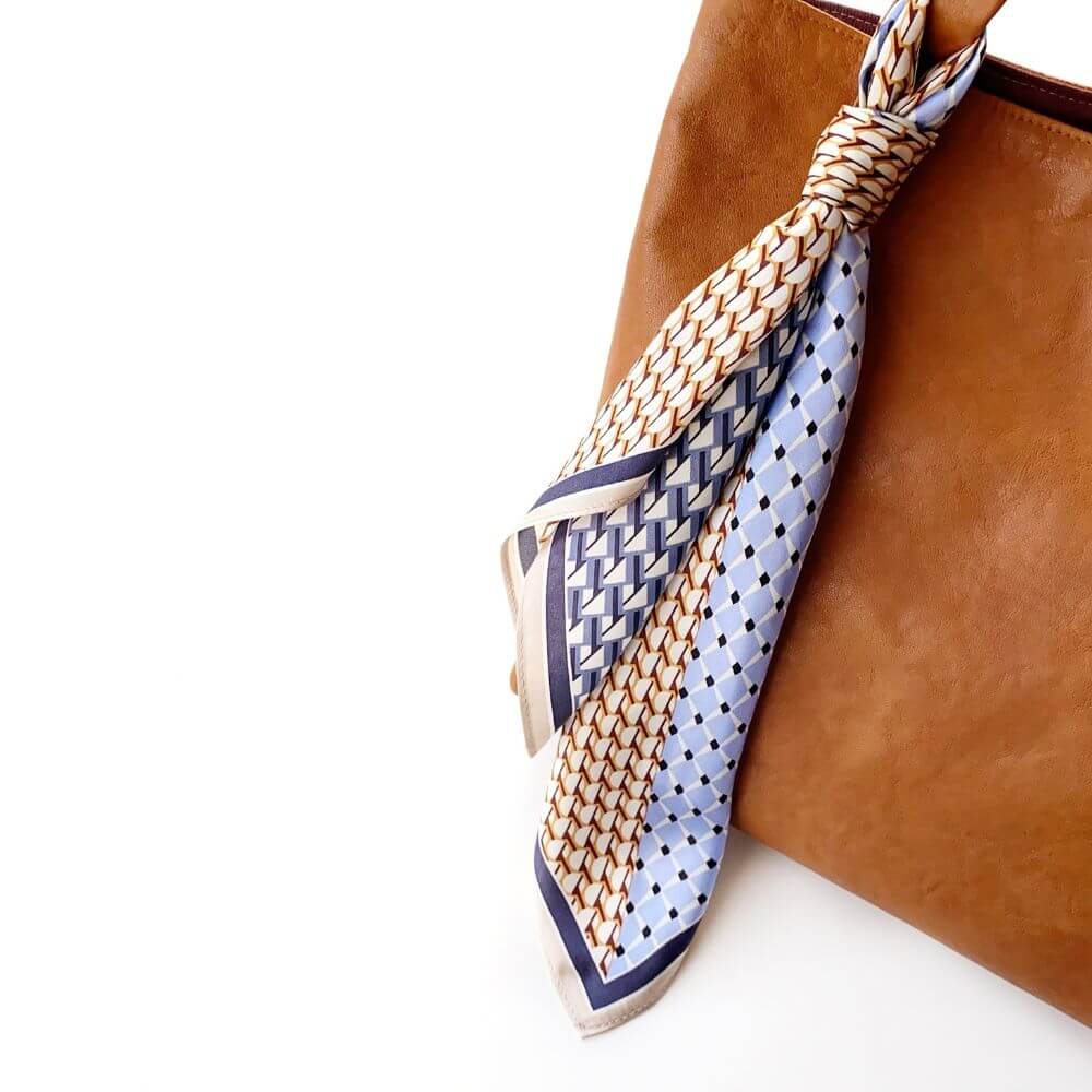 Patterned silk scarf tied around a brown leather bag on a white background