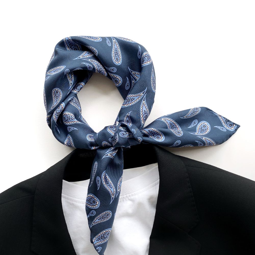 Blue paisley print mulberry silk square scarf tied into a neckerchief, placed around a black blazer and white T-shirt on a white background