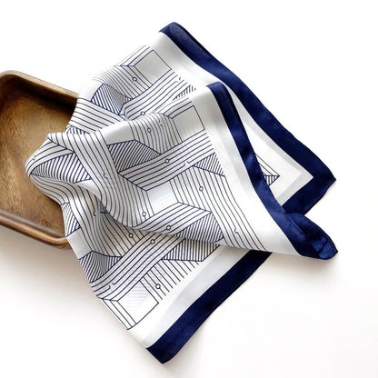 White mulberry silk scarf with a navy geometric pattern and blue border on a wooden tray.