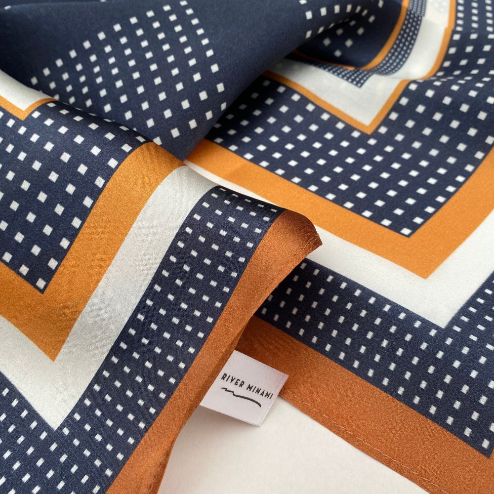 Close-up of geometric patterned mulberry silk square scarf with a visible brand tag, featuring navy, brown, and white colors.