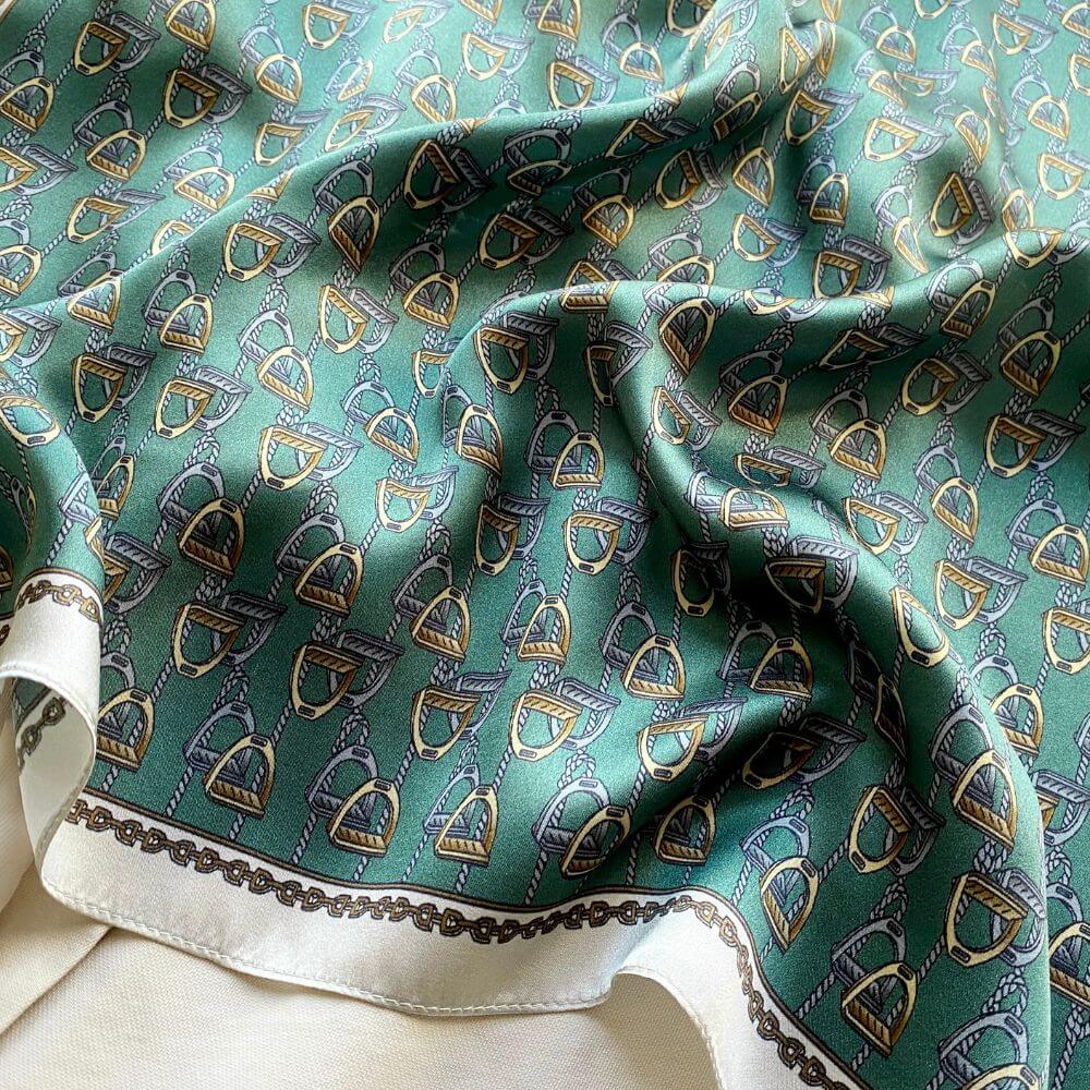 Close-up of a teal green equestrian stirrup patterned mulberry silk square scarf with gold and silver accents