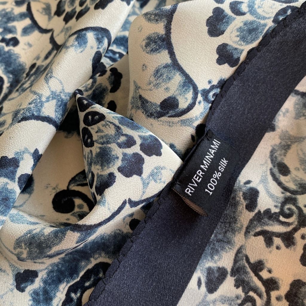 Close-up of the fabric and brand label on a mulberry silk bandana scarf showing the blue-and-white geometric floral print and a tag that reads River Minami 100% silk.