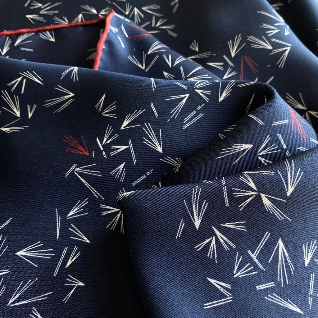 Close-up of a navy silk twill scarf with abstract white line clusters and rust-red edges.