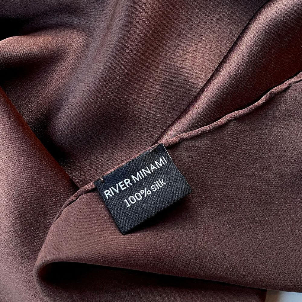 Close-up of brown silk scarf with a hand rolled hem and  a 'River Minami' label indicating 100% silk