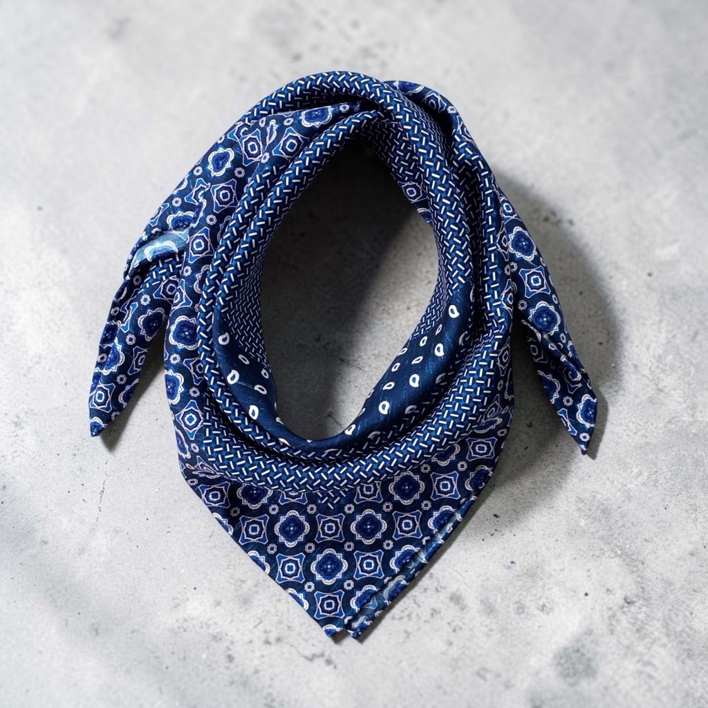 A mulberry silk square scarf in denim blue with a layered geometric, crosshatch, and paisley-inspired design placed on a light grey background.