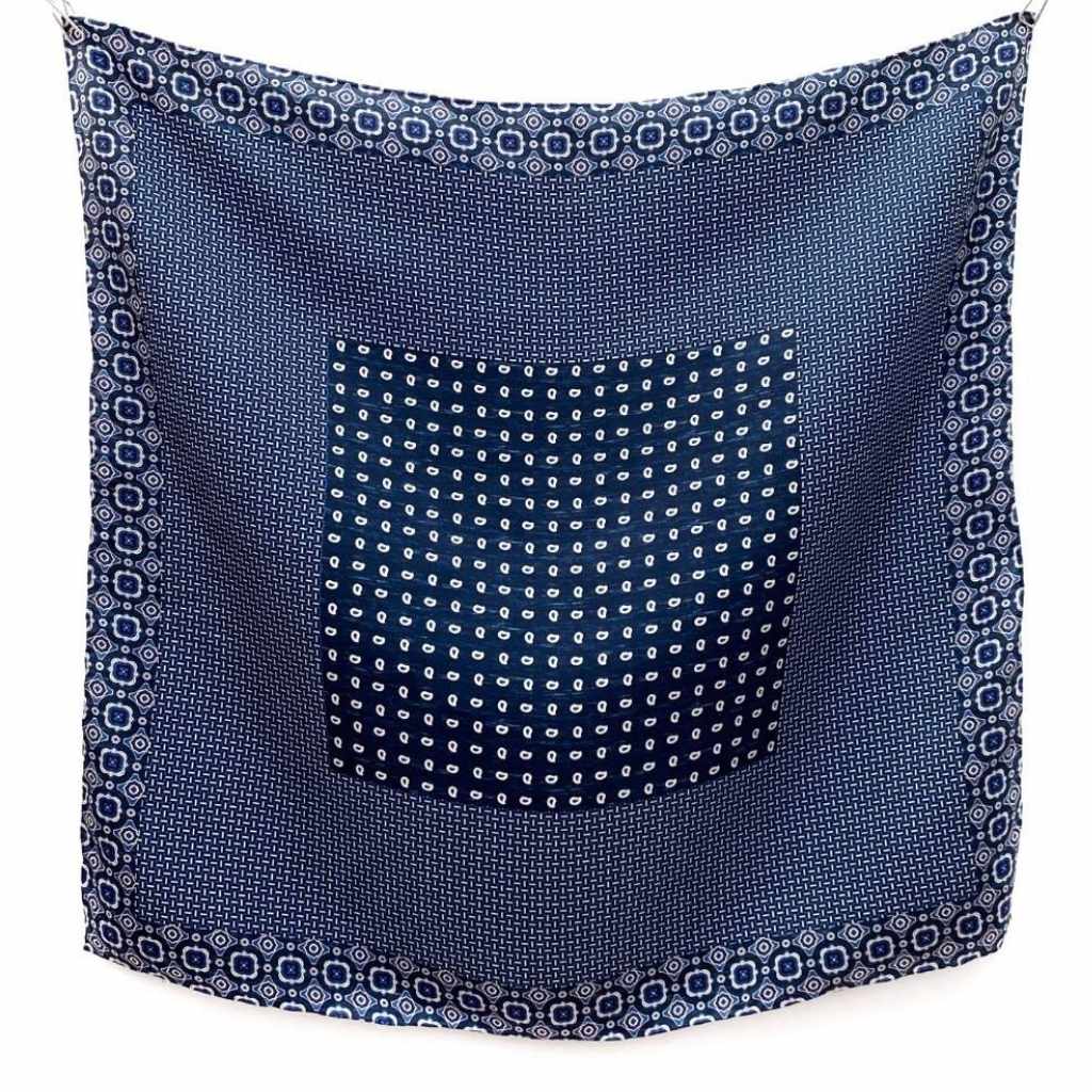 A mulberry silk square scarf in denim blue with a layered geometric, crosshatch, and paisley-inspired design hanging flat on a white wall.