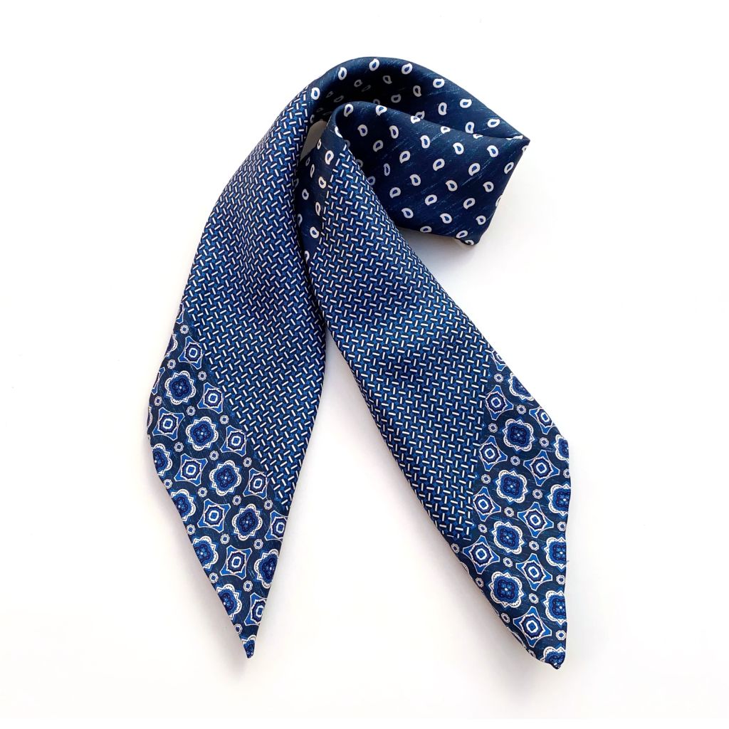 A rolled mulberry silk square scarf in denim blue with a layered geometric, crosshatch, and paisley-inspired design placed on a white background.