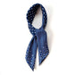 A rolled and tied mulberry silk square scarf in denim blue with a layered geometric, crosshatch, and paisley-inspired design placed on a white background.