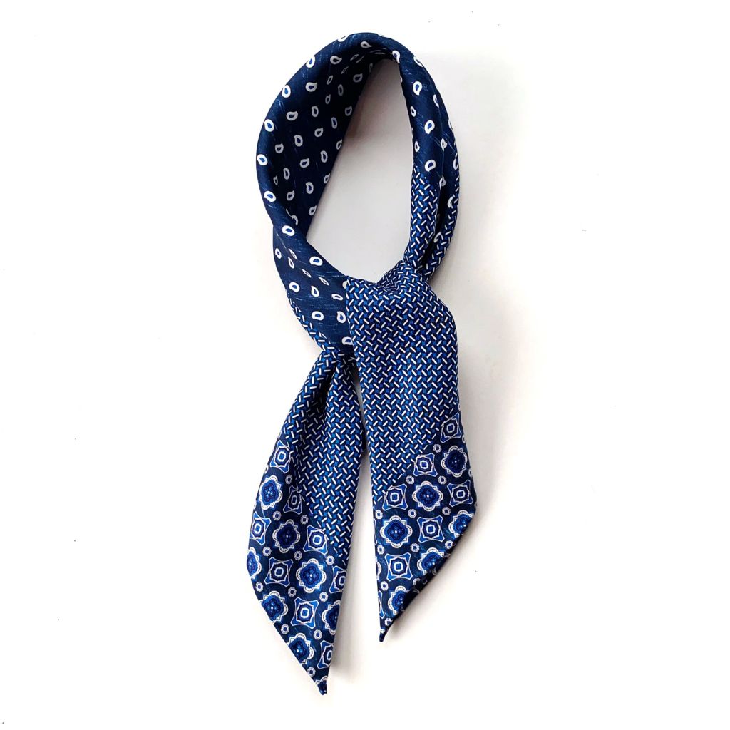 A rolled and tied mulberry silk square scarf in denim blue with a layered geometric, crosshatch, and paisley-inspired design placed on a white background.