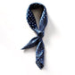 A mulberry silk square scarf in denim blue with a layered geometric, crosshatch, and paisley-inspired design tied into an ascot scarf shape, placed on a white background.