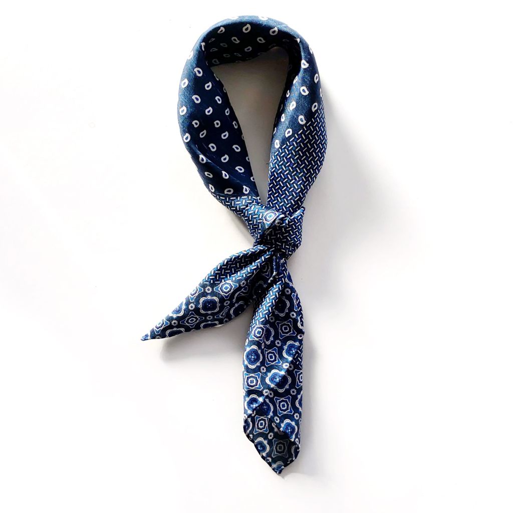 A mulberry silk square scarf in denim blue with a layered geometric, crosshatch, and paisley-inspired design tied into an ascot scarf shape, placed on a white background.
