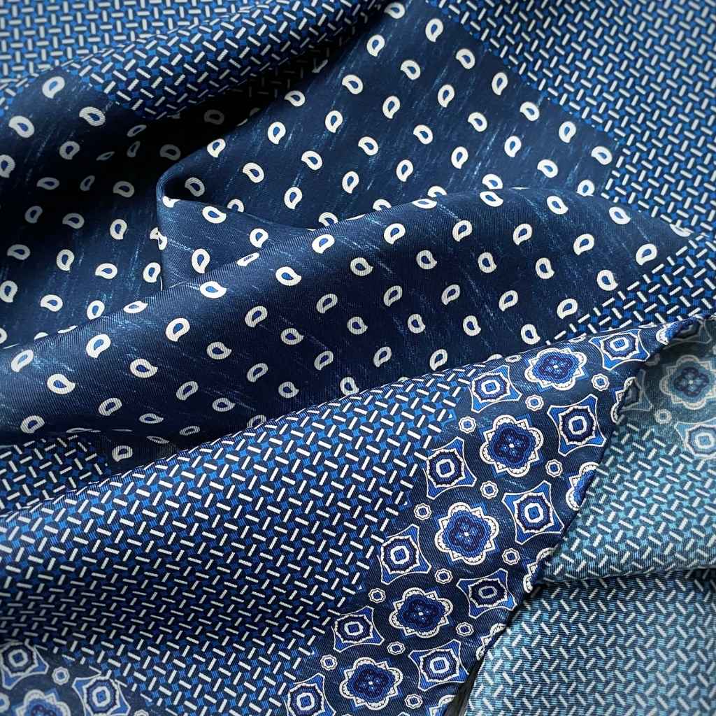 A close-up of a folded mulberry silk scarf in multiple blue patterns, including white paisley-like motifs and geometric floral designs on navy and light blue backgrounds.