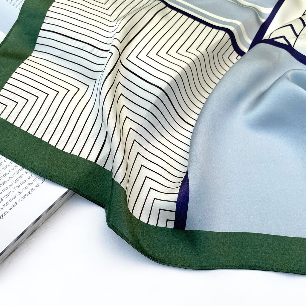 Close-up of a blue white geometric pattern mulberry silk square scarf with green edge on a white background.