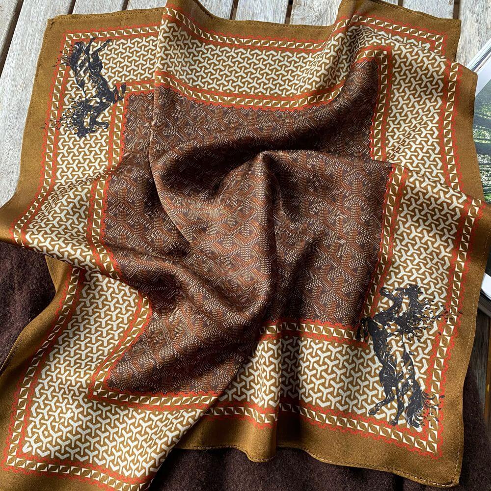 Flat lay of a brown Japanese-print pure mulberry silk square scarf featuring detailed horse designs on a wooden surface.