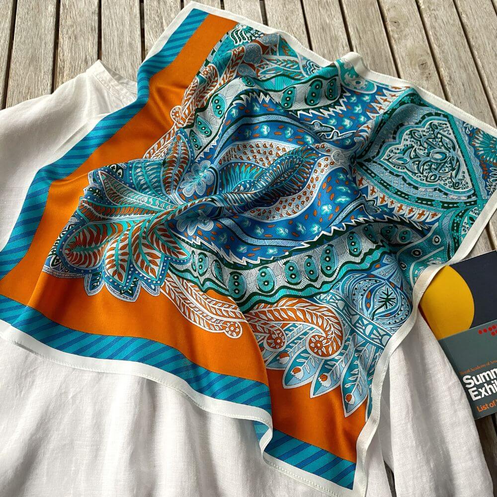 Colorful blue and orange paisley patterned mulberry silk square scarf on a white shirt lying on the table