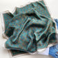 Teal green equestrian stirrup patterned mulberry silk small square scarf on a white surface with jeans in the background