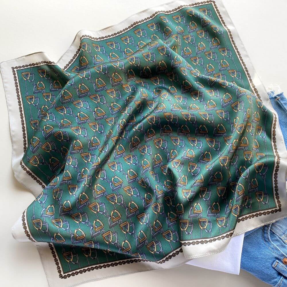 Teal green equestrian stirrup patterned mulberry silk small square scarf on a white surface with jeans in the background