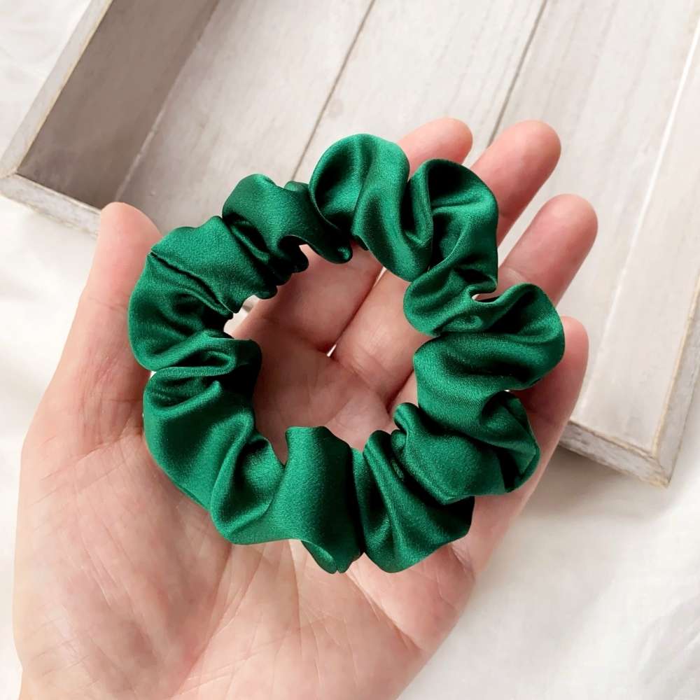 Green mulberry silk satin scrunchie held in a hand with a light wooden background