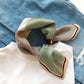 Green geometric stripes mulberry silk square scarf tied by a dark brown leather scarf clip, placed over a white shirt and blue jeans