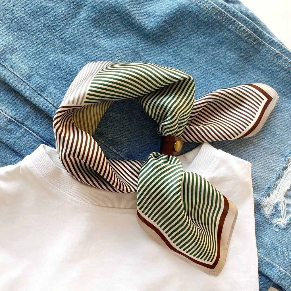 Green geometric stripes mulberry silk square scarf tied by a dark brown leather scarf clip, placed over a white shirt and blue jeans