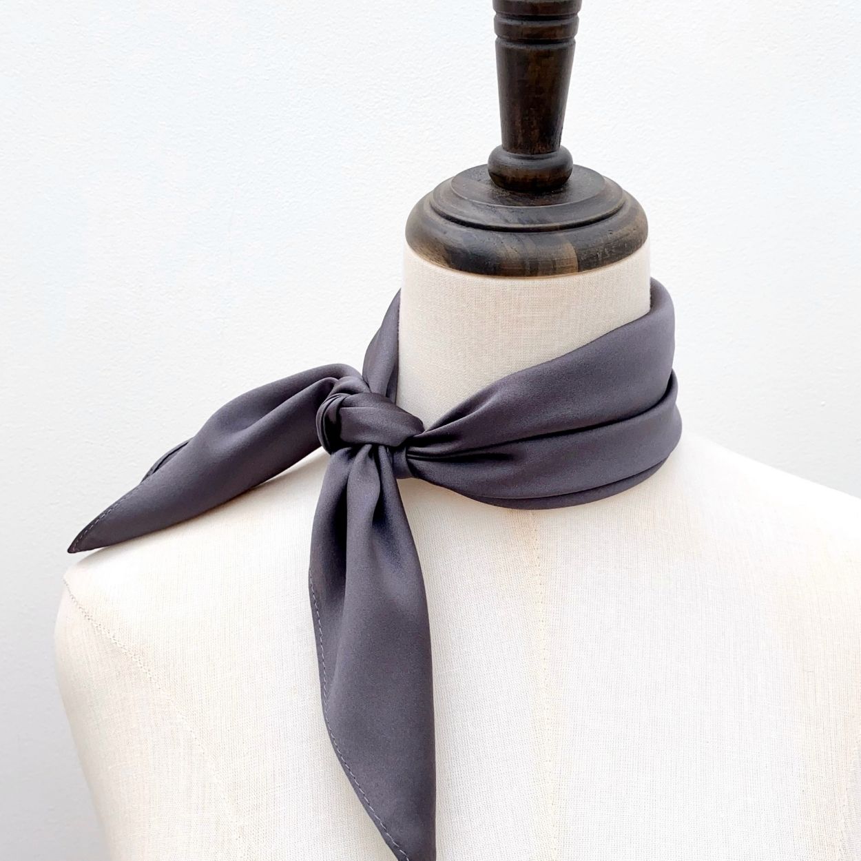 A dark gray mulberry silk square scarf tied in a knot around the neck of a mannequin, set against a plain white background.