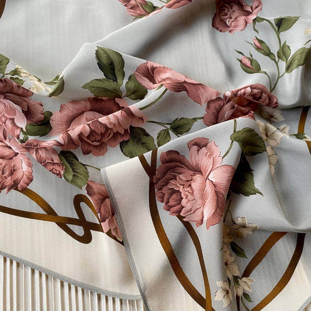 A close-up of mulberry silk crepe de chine scarf featuring a floral print with large dusty pink roses, green leaves, and gold vine outlines on a light gray background.