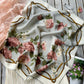 A square mulberry silk scarf with a floral print featuring dusty pink roses, green leaves, and gold wavy lines on a light beige background, styled on a rustic wooden surface.