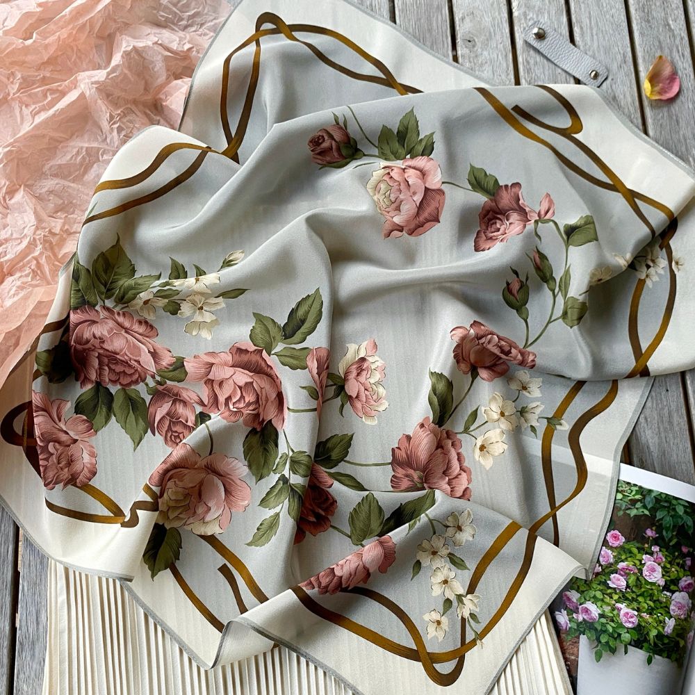 A square mulberry silk scarf with a floral print featuring dusty pink roses, green leaves, and gold wavy lines on a light beige background, styled on a rustic wooden surface.