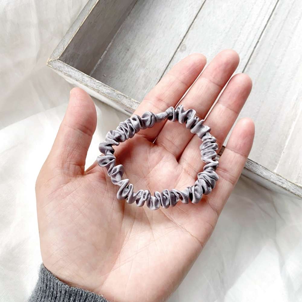 Hand holding a gray mulberry silk satin scrunchie with a light wooden frame in the background
