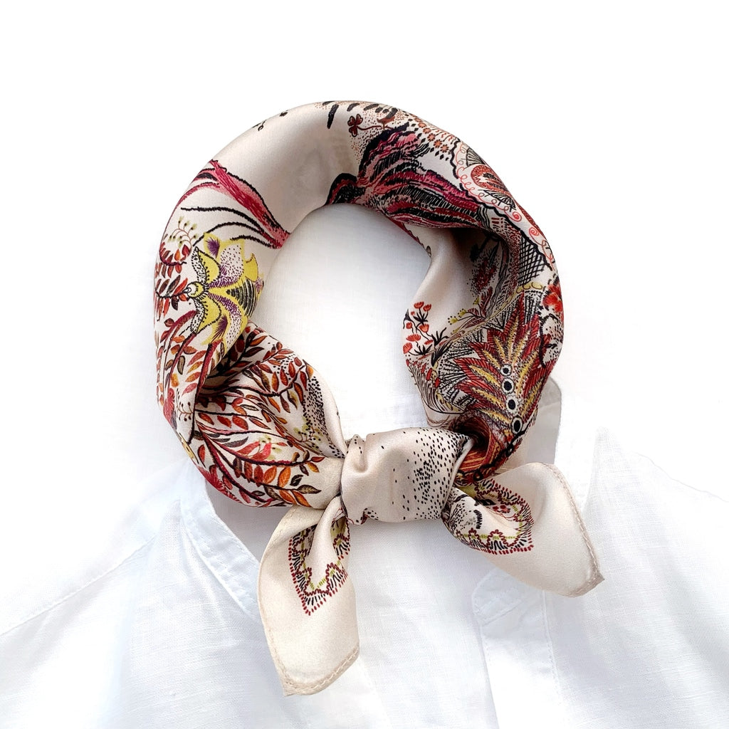 An ivory beige silk square scarf with a rusty orange floral tree of life print, displayed on a white shirt collar.