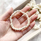 Hand holding an ivory cream mulberry silk satin skinny scrunchie with more bracelets in the background.