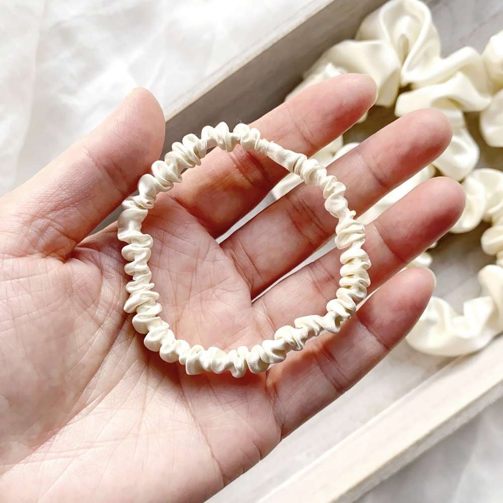 Hand holding an ivory cream mulberry silk satin skinny scrunchie with more bracelets in the background.