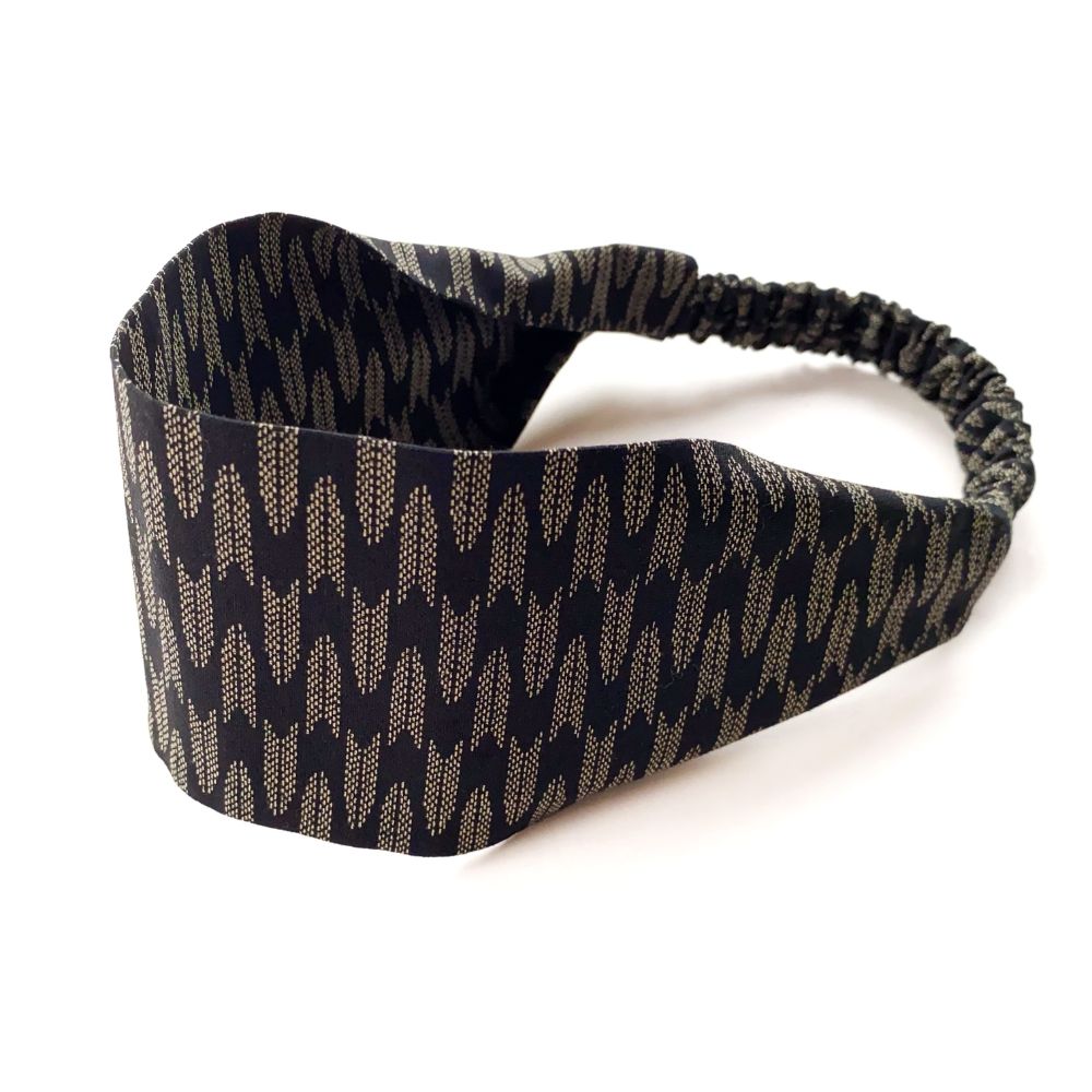 A wide cotton fabric headband with an elastic back, featuring a repeating arrow pattern in beige and black.
