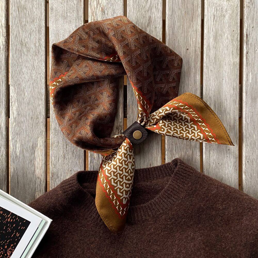 Brown Japanese patterned mulberry silk bandana scarf neatly tied with a leather scarf clip placed around a brown sweater, set against a rustic wooden background.