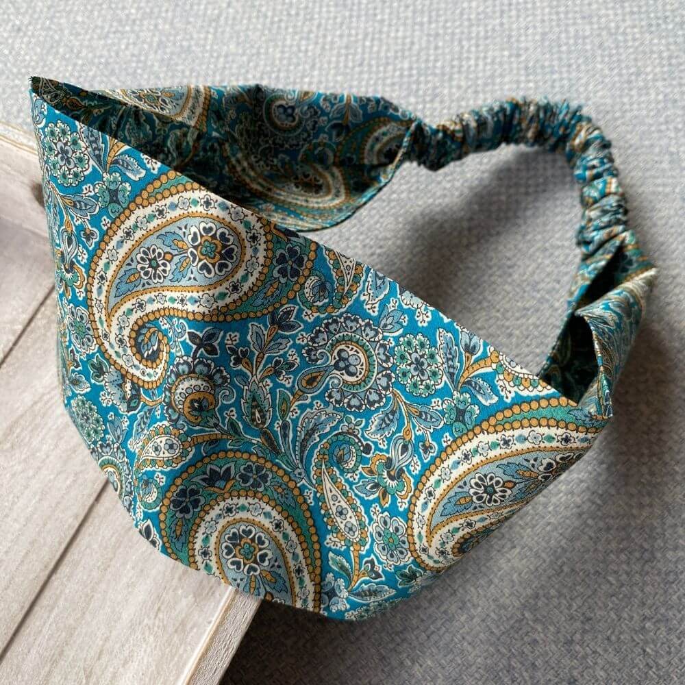 Liberty Lee Manor cotton headband with a wide flat front and elasticated back, shown in a relaxed angled view highlighting the teal and mustard paisley print.