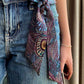 A close-up of a person wearing blue denim shorts and a patterned liberty london silk skinny scarf tied around the waist. The scarf features an intricate, multicolored abstract design with purple, teal, gold, and black elements.