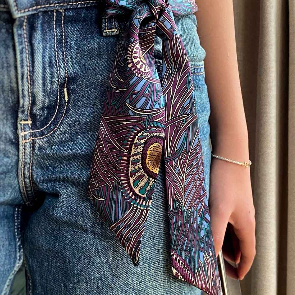 A close-up of a person wearing blue denim shorts and a patterned liberty london silk skinny scarf tied around the waist. The scarf features an intricate, multicolored abstract design with purple, teal, gold, and black elements.