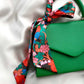 A green textured leather handbag with a vibrant floral liberty london silk twilly scarf tied in a knot and draped over the handle, set against a white satin background.