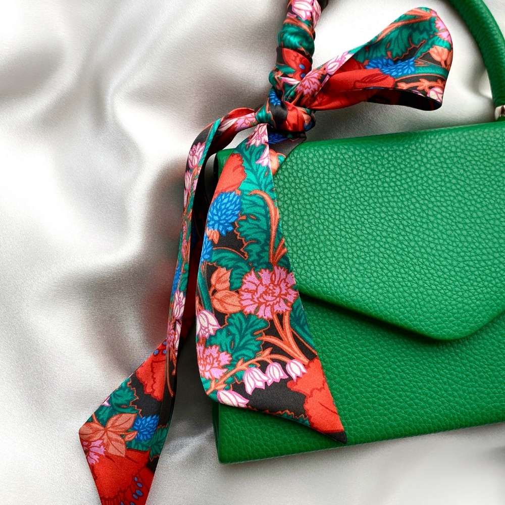 A green textured leather handbag with a vibrant floral liberty london silk twilly scarf tied in a knot and draped over the handle, set against a white satin background.