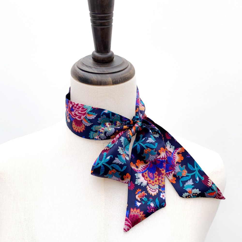 A patterned liberty london silk skinny scarf with a dark navy base and colorful floral paisley motifs, styled on a mannequin neck.