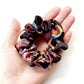 A close-up of a hand holding a liberty silik satin hair scrunchie with a colorful abstract pattern on a white background.