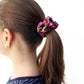 A close-up side view of a person with long brown hair tied back in a ponytail using a colorful liberty silk scrunchie against a plain white background.