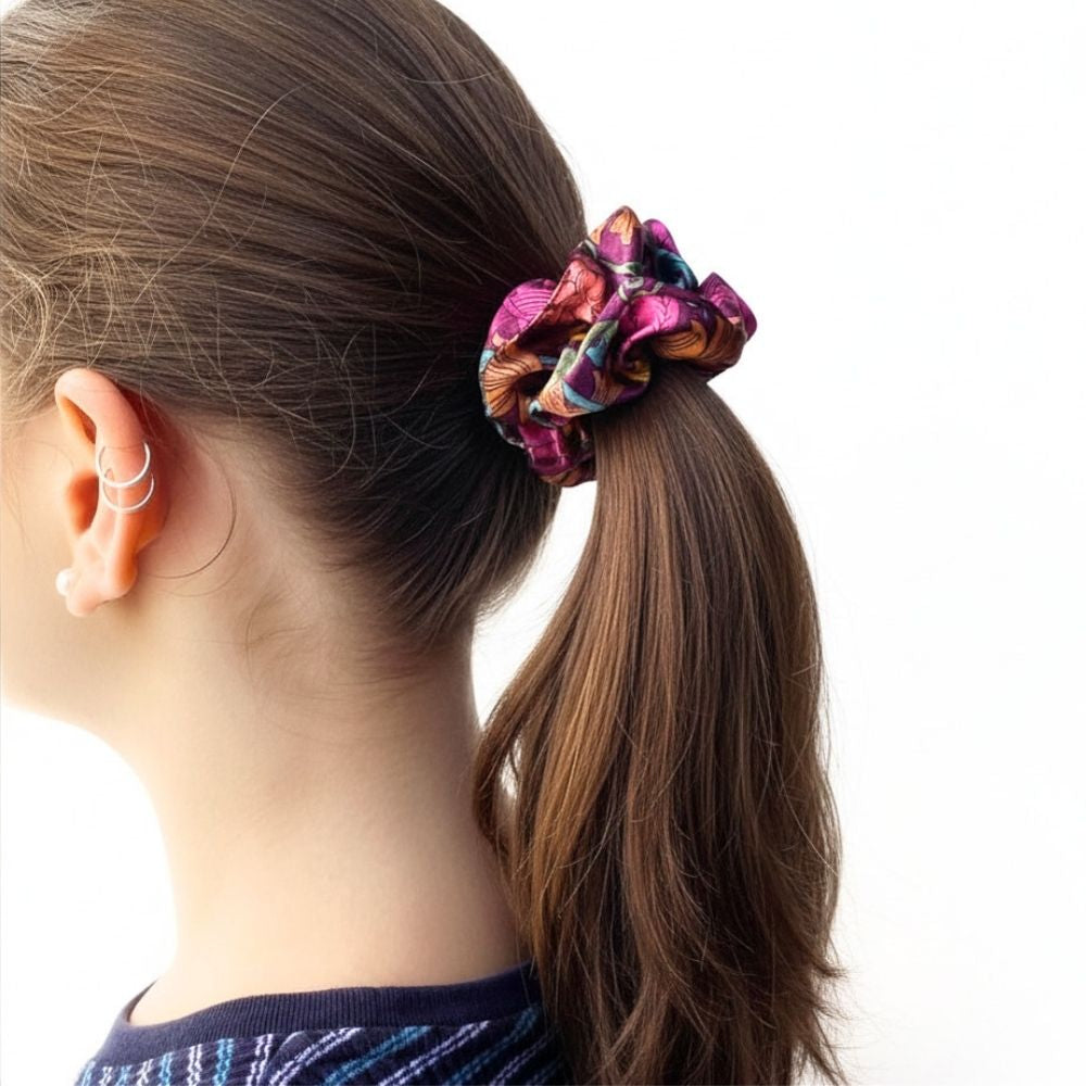 A close-up side view of a person with long brown hair tied back in a ponytail using a colorful liberty silk scrunchie against a plain white background.